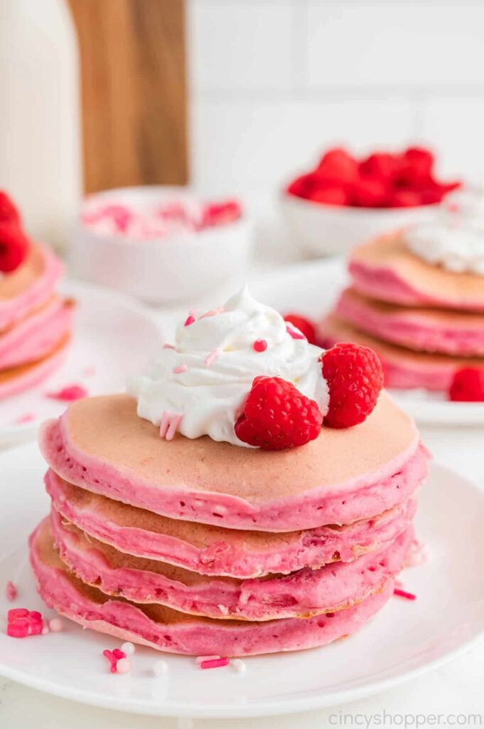 white plate with pink pancake stack with whipped cream and raspberries